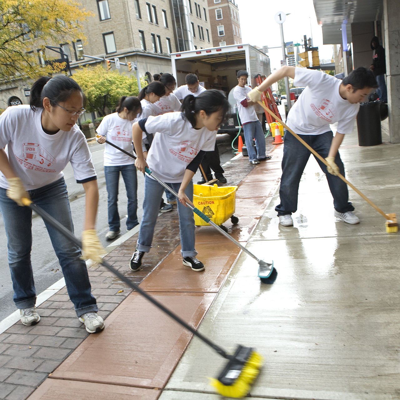Several young adults use brooms to scrub a city sidewalk.