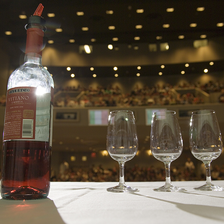 An open bottle of wine and three empty wine glasses sit at the font of a full auditorium.