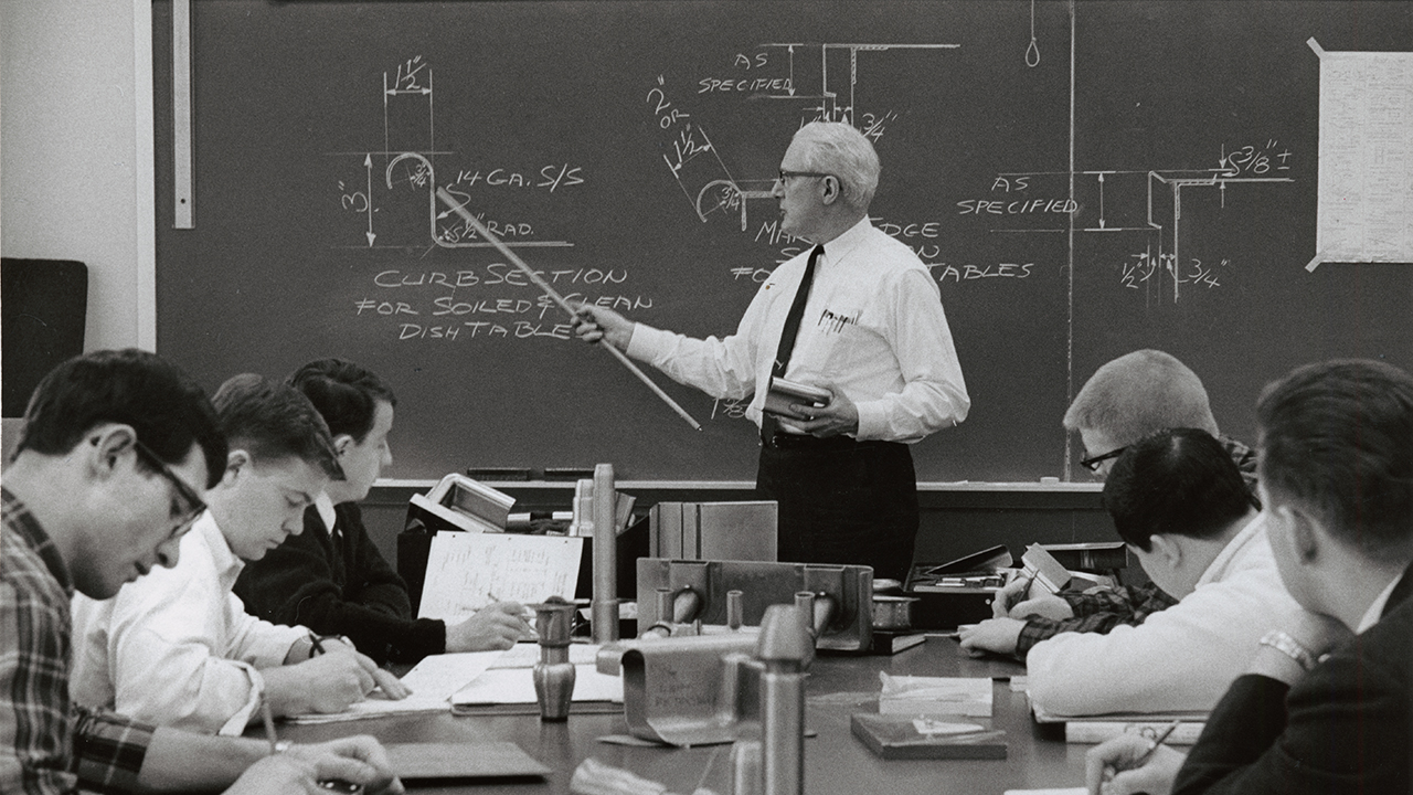 A teacher holding a pointer in front of a blackboard,  6 students at a table taking notes.