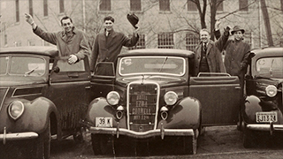 Historical photo of men standing by cars with '14th Annual Hotel Ezra Cornell May 12' painted on one car grid