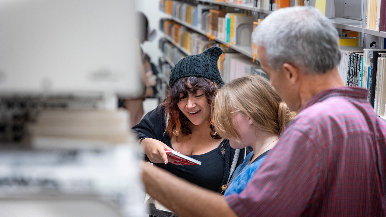Three people investigate a book within the library stacks.